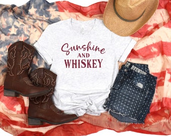 May include: A white t-shirt with the text "Sunshine and Whiskey" in maroon lettering. The shirt is laid out on a red, white, and blue patterned fabric with a pair of brown cowboy boots, a straw hat, and denim shorts with white polka dots.