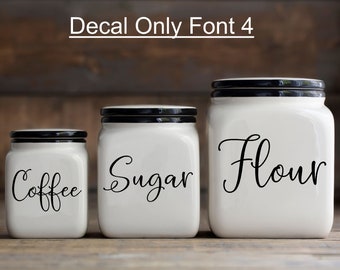 May include: Three white ceramic canisters with black lids and black vinyl lettering. The canisters are labeled "Coffee", "Sugar", and "Flour" in a cursive font.