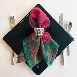 May include: A red and green plaid napkin with a white and gold beaded napkin ring on a black square plate. The napkin is folded into a triangle shape and the ring is placed at the top of the triangle.