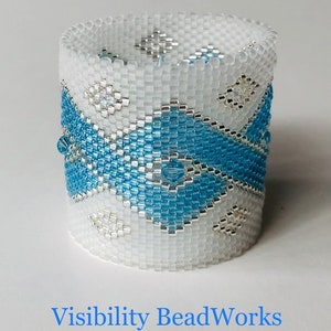 May include: A white and blue beaded napkin ring with a geometric pattern. The ring is made with small, square beads and features a blue and white design.