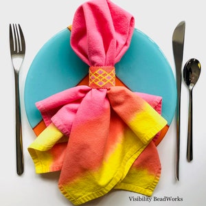 May include: A pink and yellow ombre napkin is folded and placed on a blue and orange plate. A beaded napkin ring is on the napkin. The napkin is set with a fork and knife on a white background.