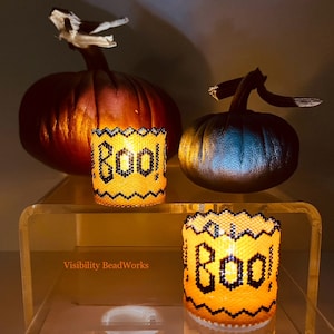 May include: Two orange and black beaded candle holders with the word "BOO!" on them. The candle holders are sitting on a clear acrylic surface with two pumpkins behind them.