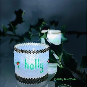 May include: A white beaded candle holder with the name "Holly" in green letters, accented with red berries and green leaves. The top and bottom edges are trimmed with a black and gold pattern. A second candle holder is visible in the background.