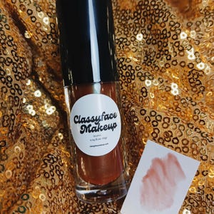 May include: A clear glass bottle of Classyface Makeup lip gloss with a black cap, resting on a gold sequined surface. A white label with the brand name is visible. A swatch of the lip gloss is on a small white card.