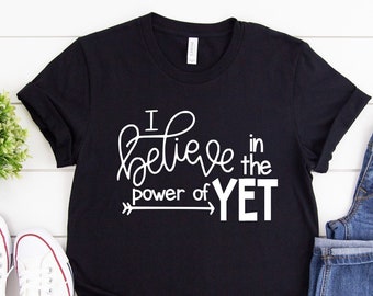 Puede incluir: Camiseta negra con texto blanco que dice "I believe in the power of YET" con una flecha apuntando hacia la derecha. La camiseta está colocada sobre una superficie de madera blanca con un par de zapatillas blancas y unos vaqueros azules.