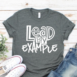 May include: A dark gray t-shirt with the text "Lead by Example" in white lettering. The shirt is laid flat on a white wooden surface with a pair of white sneakers and blue jeans.