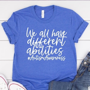 May include: A royal blue t-shirt with white text that reads "We all have different abilities #AutismAwareness". The shirt is laid flat on a white wooden surface with a pair of white sneakers and blue jeans.