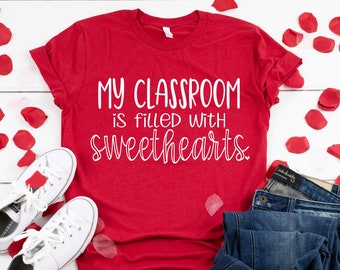 Puede incluir: Camiseta roja con el texto "My classroom is filled with sweethearts." sobre un fondo blanco con pétalos de rosa roja, un par de zapatillas blancas y unos vaqueros azules.