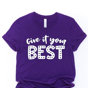 May include: Purple t-shirt with white text that reads "Give it your BEST" with white polka dots around the letters.