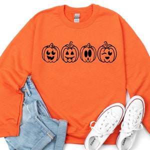 May include: Orange sweatshirt with a black graphic of four pumpkins with different facial expressions. The sweatshirt is laying on a white surface with a pair of white sneakers.