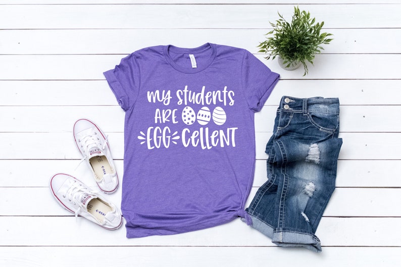 Może przedstawiać: Fioletowa koszulka z białym napisem "my students ARE EGG-CELLENT". Na zdjęciu widoczne są niebieskie dżinsy i białe trampki. Widoczna jest r&oacute;wnież mała roślina doniczkowa.