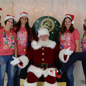 Puede incluir: Un grupo de mujeres con camisas rojas con luces de Navidad y el texto "The Bells Still Ring For Me" posan con Pap&aacute; Noel. Las mujeres llevan gorros de Pap&aacute; Noel y pantalones vaqueros. Pap&aacute; Noel lleva un traje rojo con ribetes blancos y una barba blanca.
