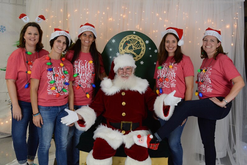 Puede incluir: Un grupo de mujeres con camisas rojas con luces de Navidad y el texto "The Bells Still Ring For Me" posan con Pap&aacute; Noel. Las mujeres llevan gorros de Pap&aacute; Noel y pantalones vaqueros. Pap&aacute; Noel lleva un traje rojo con ribetes blancos y una barba blanca.