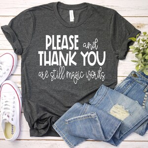May include: A dark gray t-shirt with the text "PLEASE and THANK YOU are still magic words" printed in white.