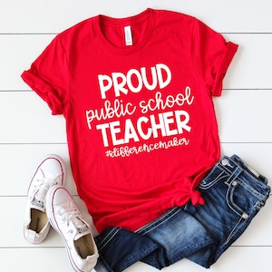 May include: Red t-shirt with white text that reads "Proud public school teacher #differencemaker". The shirt is worn with blue jeans and white trainers.