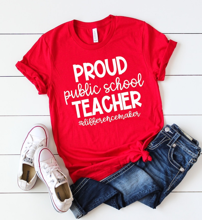 May include: Red t-shirt with white text that reads "Proud public school teacher #differencemaker". The shirt is worn with blue jeans and white trainers.