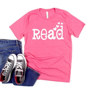 May include: Pink t-shirt with white text that says "Read" with small white hearts around the letters.