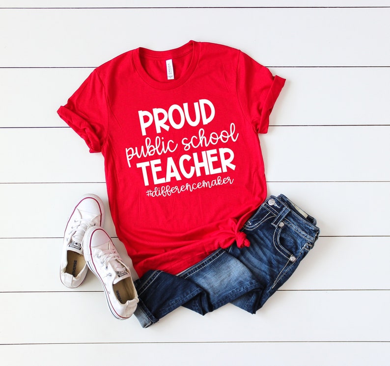 May include: Red t-shirt with white text that reads "Proud public school teacher #differencemaker". The shirt is worn with blue jeans and white trainers.