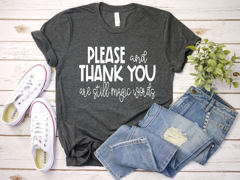 May include: A dark gray t-shirt with white text that reads "PLEASE and THANK YOU are still magic words". The shirt is laid flat on a white wooden surface with a pair of white sneakers and blue jeans.