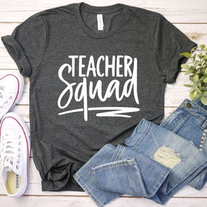 Może przedstawiać: Szary t-shirt z napisem "Teacher Squad" w białym kolorze. Koszulka jest złożona i leży na białym drewnianym tle z parą białych but&oacute;w sportowych i niebieskimi dżinsami.