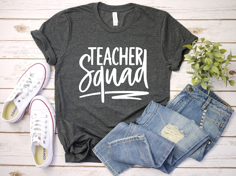Może przedstawiać: Szary t-shirt z napisem "Teacher Squad" w białym kolorze. Koszulka jest złożona i leży na białym drewnianym tle z parą białych but&oacute;w sportowych i niebieskimi dżinsami.