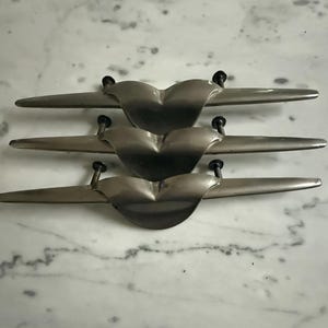 May include: Three silver-toned metal drawer pulls arranged horizontally. Each pull features a central curved, wing-like shape that tapers to pointed ends, with two small black mounting screws attached to the back of each piece for installation.