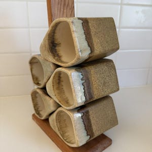 Puede incluir: Un juego de seis tazas de cerámica en un soporte de madera. Las tazas son rectangulares con bordes redondeados, con un exterior moteado de color beige y un interior esmaltado blanco y marrón. El soporte de madera tiene un acabado natural.