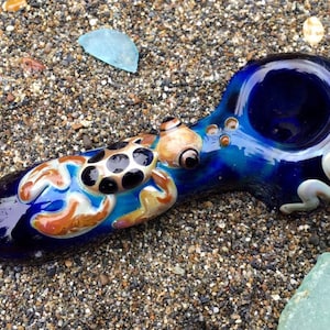 May include: A hand-blown glass pipe with a deep blue hue, featuring an octopus design in shades of tan, white, and black. The pipe has a bowl and a stem, with a swirling white and tan accent. The pipe is resting on a sandy surface.