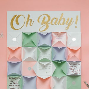 May include: A white board with the words "Oh Baby!" in gold glitter letters. The board is decorated with colorful paper envelopes, each with a handwritten message inside. The envelopes are arranged in a geometric pattern.