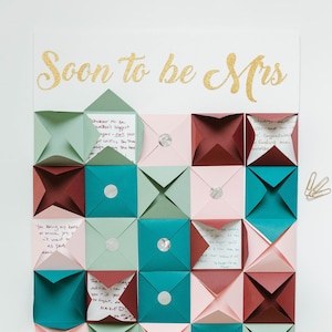 Puede incluir: Una pizarra blanca con el texto "Soon to be Mrs" en letras doradas con purpurina. La pizarra está decorada con una cuadrícula de 16 sobres de papel doblados en tonos rosa, verde y burdeos. Cada sobre tiene una pequeña pegatina redonda plateada en la parte delantera.