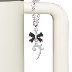 May include: A silver-toned charm with a black bow and a treble clef hangs from a white mug handle. The charm is attached to a chain. The mug is white with a silver top.