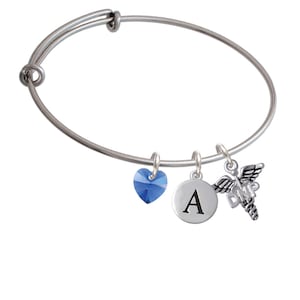 May include: A silver bangle bracelet with a blue crystal heart charm, a silver charm with the letter A, and a silver charm of a caduceus with the letters DNP.