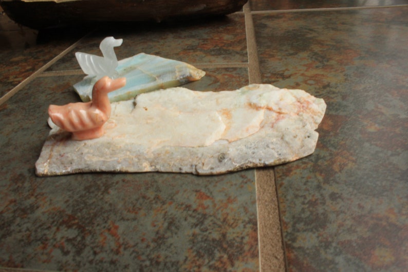May include: A pink and white carved stone duck figurine sits on a larger piece of white and brown stone. The stone is textured and has a natural, rough look.
