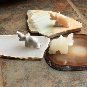 May include: Three small elephant figurines made of white, grey, and beige stone, each sitting on a different agate slice. The agate slices are in shades of brown, white, and grey.