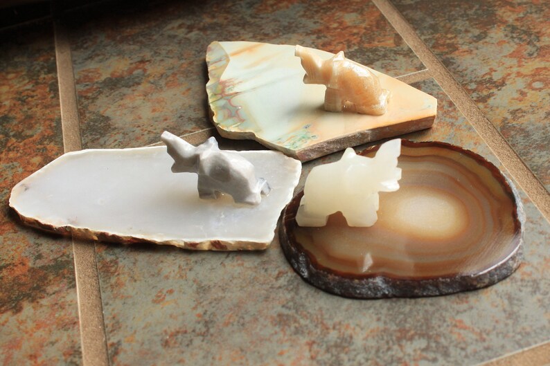 May include: Three small elephant figurines made of white, grey, and beige stone, each sitting on a different agate slice. The agate slices are in shades of brown, white, and grey.