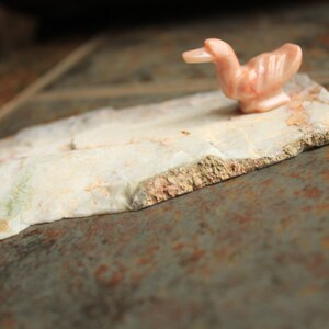 May include: A small, pink, carved duck figurine sits on a piece of white, marbled stone. The stone has a rough, textured surface.