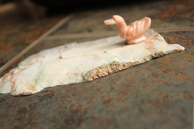 May include: A small, pink, carved duck figurine sits on a piece of white, marbled stone. The stone has a rough, textured surface.