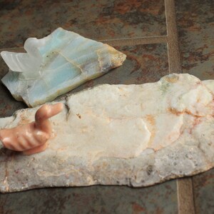 May include: A pink carved duck figurine sits on a light brown and white stone slab. A light blue and white stone slab with a crystal formation is in the background.