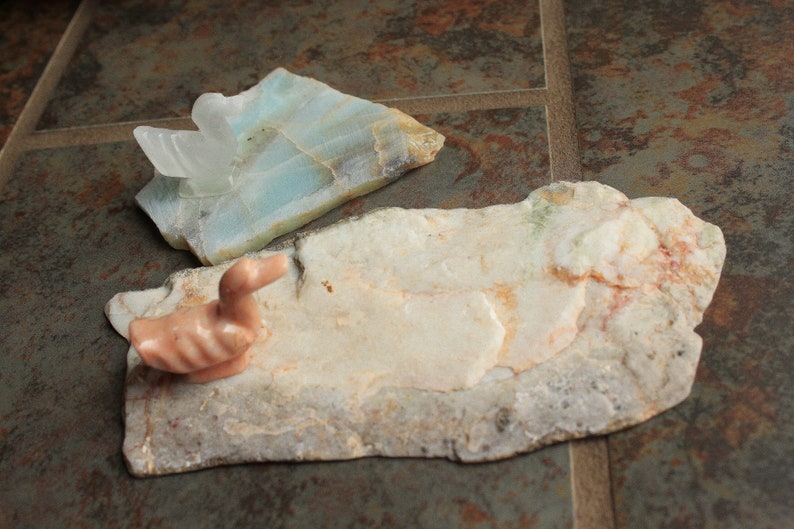 May include: A pink carved duck figurine sits on a light brown and white stone slab. A light blue and white stone slab with a crystal formation is in the background.