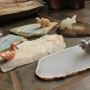 May include: Five small, carved animal figurines on various natural stone bases. The figurines include a white elephant, a grey rhinoceros, a pink duck, a white swan, and a tan dinosaur. The bases are made of agate, jasper, and other natural stones.