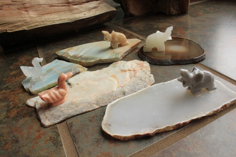 May include: Five small, carved animal figurines on various natural stone bases. The figurines include a white elephant, a grey rhinoceros, a pink duck, a white swan, and a tan dinosaur. The bases are made of agate, jasper, and other natural stones.