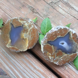 May include: Two circular agate geodes sliced in half, revealing a smooth, translucent periwinkle blue center surrounded by a thick, textured, earthy brown outer crust with visible cracks and mineral deposits throughout the stone surfaces.