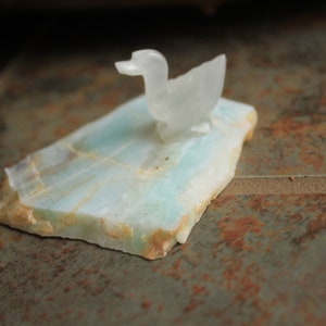 May include: A white carved duck figurine sits on a light blue and green stone base.