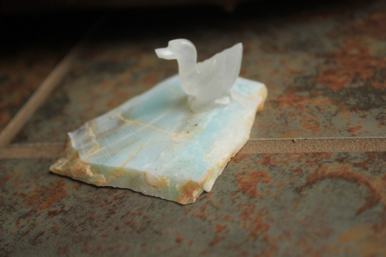 May include: A white carved duck figurine sits on a light blue and green stone base.