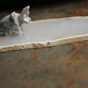 May include: A small, grey, carved elephant figurine sits on a white agate coaster. The coaster has a natural, irregular shape and a smooth, polished surface.