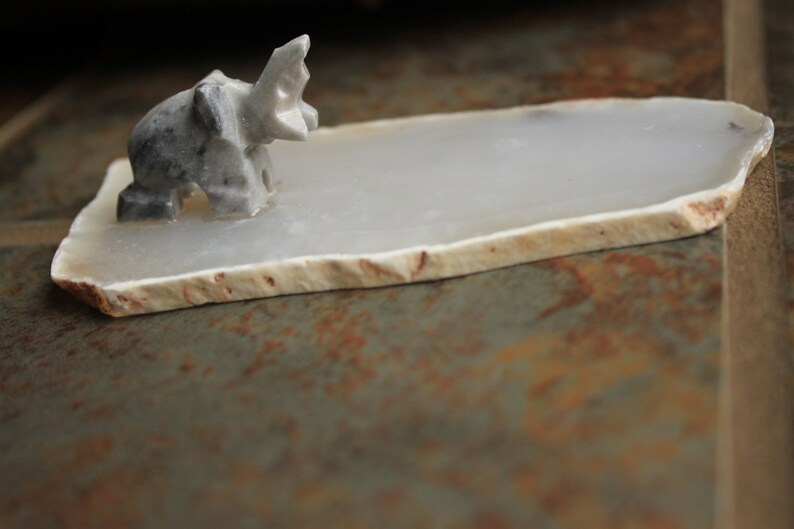 May include: A small, grey, carved elephant figurine sits on a white agate coaster. The coaster has a natural, irregular shape and a smooth, polished surface.