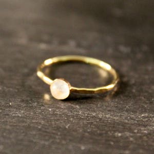 May include: A delicate gold ring with a small, round, pale gemstone. The ring has a hammered texture on the band and sits on a dark, textured surface. The gemstone appears to be a light, neutral color.