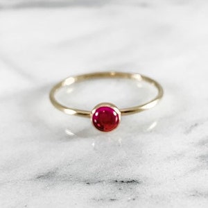 Może przedstawiać: Prosty złoty pierścionek z małym, okrągłym, czerwonym kamieniem szlachetnym.