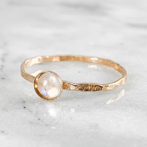 May include: A gold ring with a round, iridescent moonstone. The band has a hammered texture. The ring is set against a white marble surface.