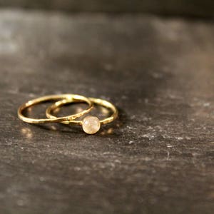 May include: Two gold rings, one with a small, round, light-colored stone, rest on a dark, textured surface. The rings have a simple, elegant design, perfect for everyday wear or special occasions.